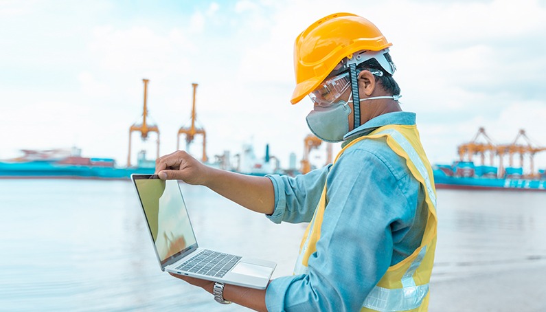 Construction worker opens a laptop outdoors, with oil rigs in the background